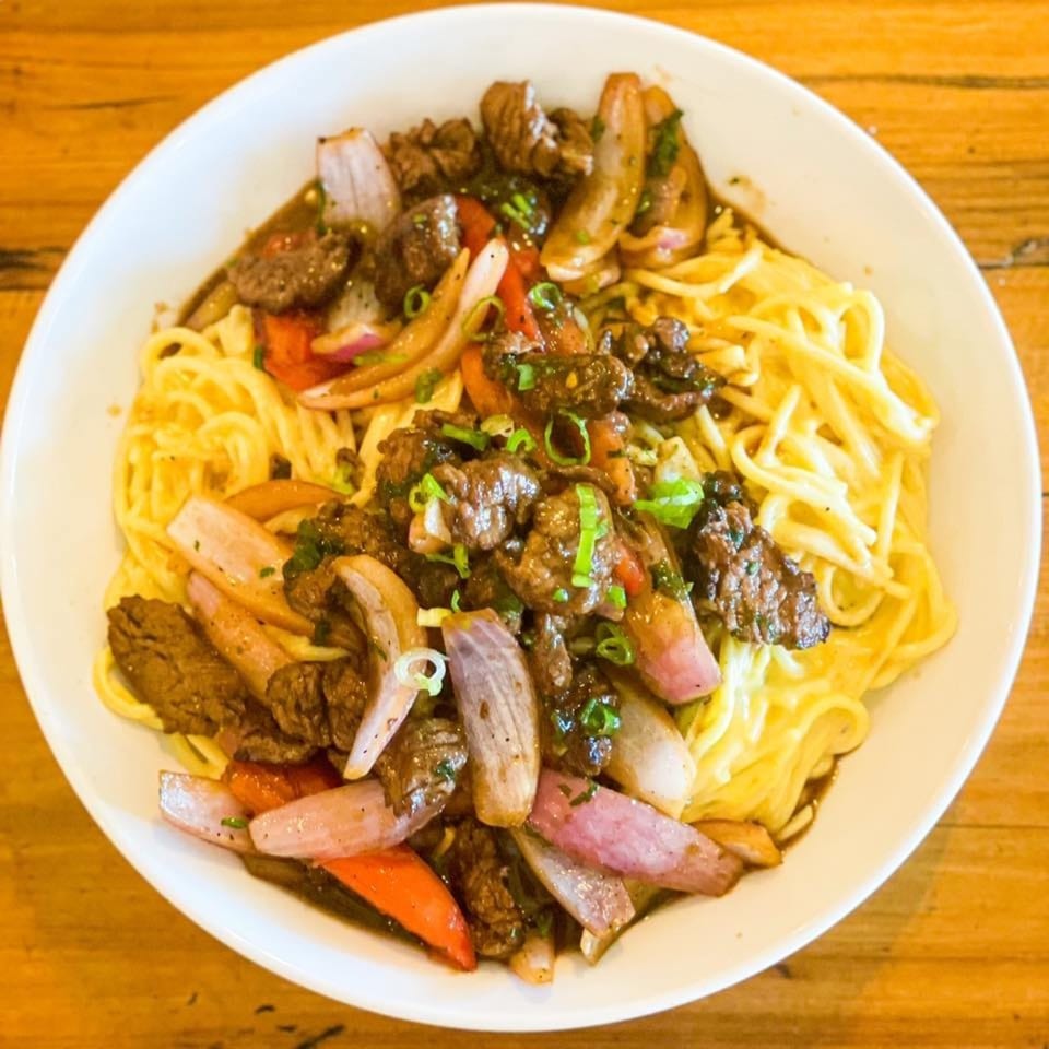 Peruvian Lomo Saltado with beef, red onions, tomatoes, and noodles in a white bowl.