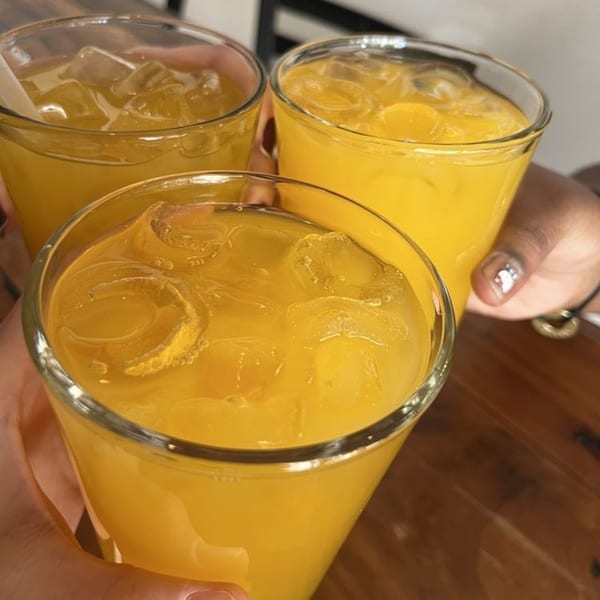 Three hands holding glasses of bright yellow Peruvian chicha morada with ice cubes.