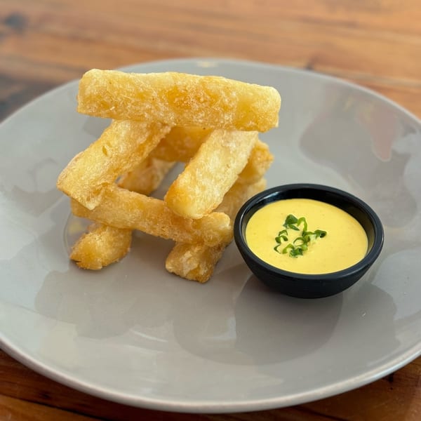 Golden yuca fries stacked on a gray plate next to a black bowl of yellow dipping sauce.