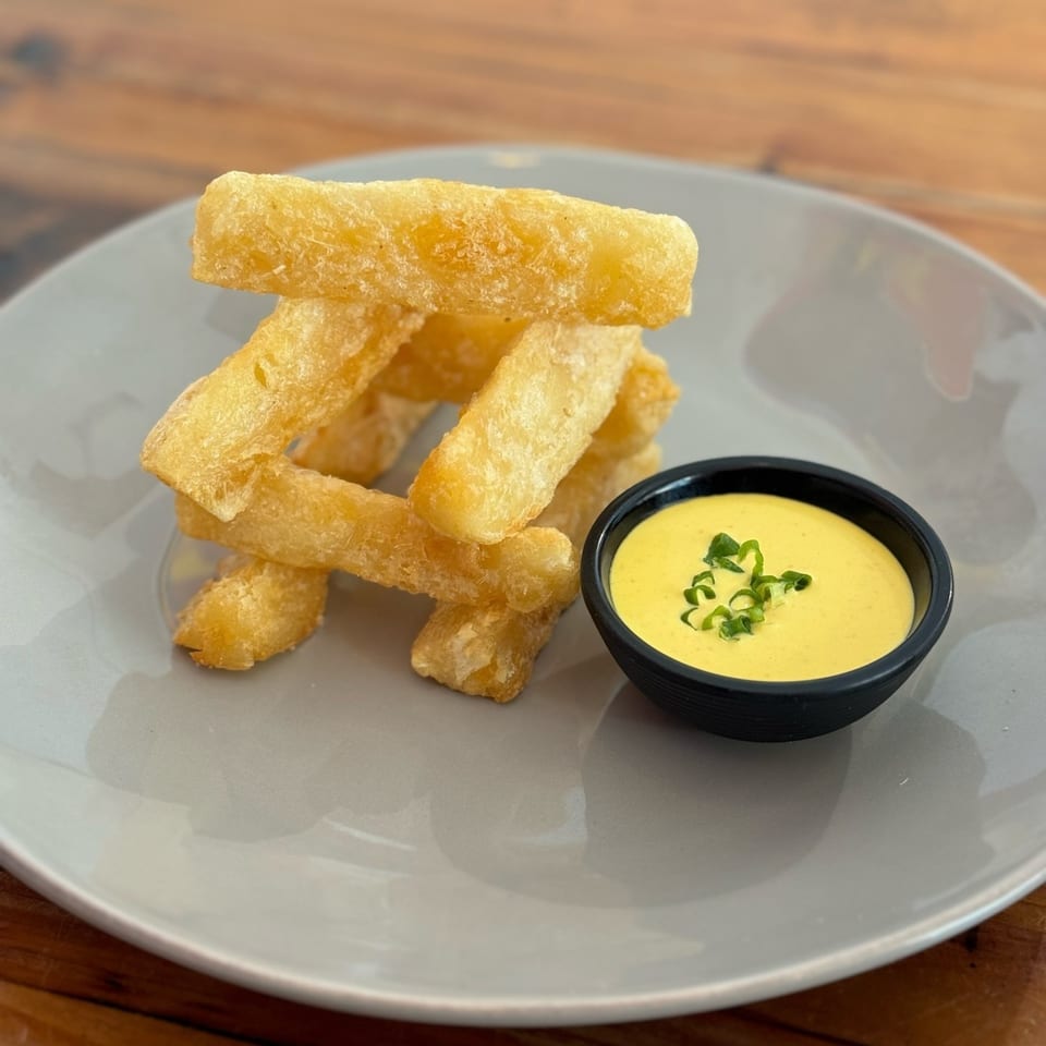 Golden yuca fries stacked on a gray plate next to a black bowl of yellow dipping sauce.